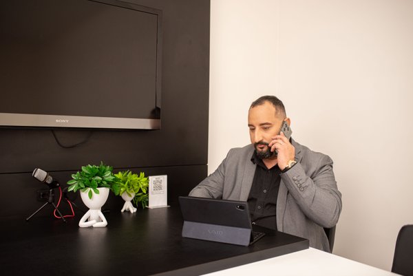 Gabriel Carrara - homem de porte grande, moreno, barba e calvo. Segurando telefone celular no ouvido e olhando no laptop. Usa terno cinca e camissão social preta.