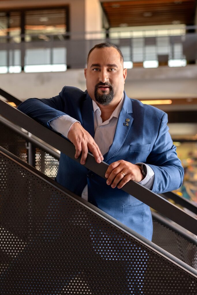 Gabriel Carrara de terno azul, camisa social branca por baixo do paletó. Em pé, de lado numa escada de ferro, cor marrom.